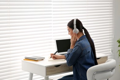 Photo of Online translation course. Student in headphones writing near laptop indoors, space for text