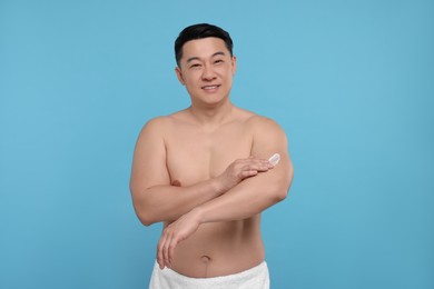 Photo of Handsome man applying body cream onto his arm on light blue background