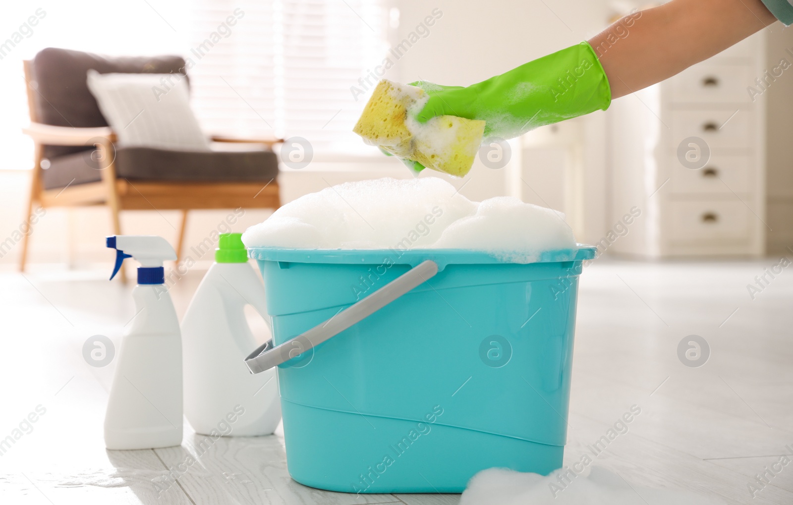 Photo of Woman holding sponge with foam over bucket indoors, closeup. Cleaning supplies