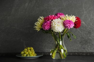 Photo of Bouquet of beautiful Dahlia flowers in vase and plate with grapes on black table near grey wall