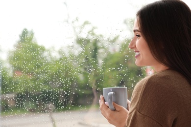 Photo of Beautiful woman with cup of coffee smiling near window indoors on rainy day