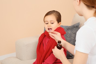 Mother giving cough syrup to her daughter from measuring cup on sofa indoors