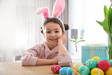 Photo of Cute little girl with bunny ears headband and painted Easter eggs sitting at table in room
