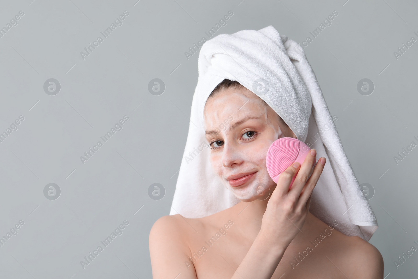 Photo of Young woman washing face with brush and cleansing foam on grey background. Space for text