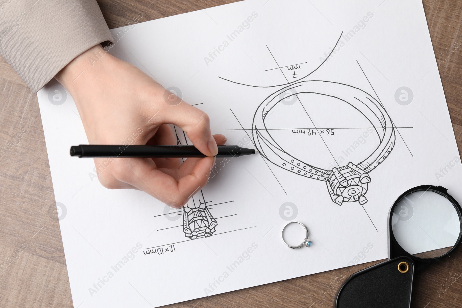 Photo of Jeweler drawing sketch of elegant ring on paper at wooden table, closeup