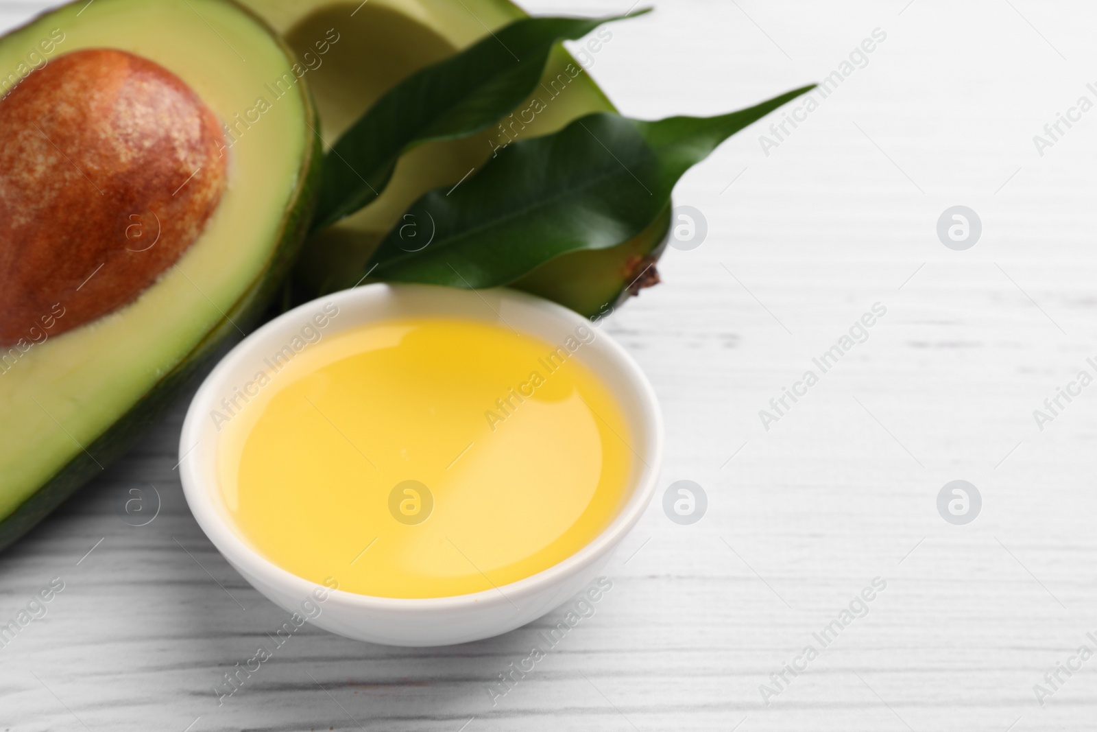 Photo of Cooking oil in bowl and fresh avocados on white wooden table, closeup. Space for text