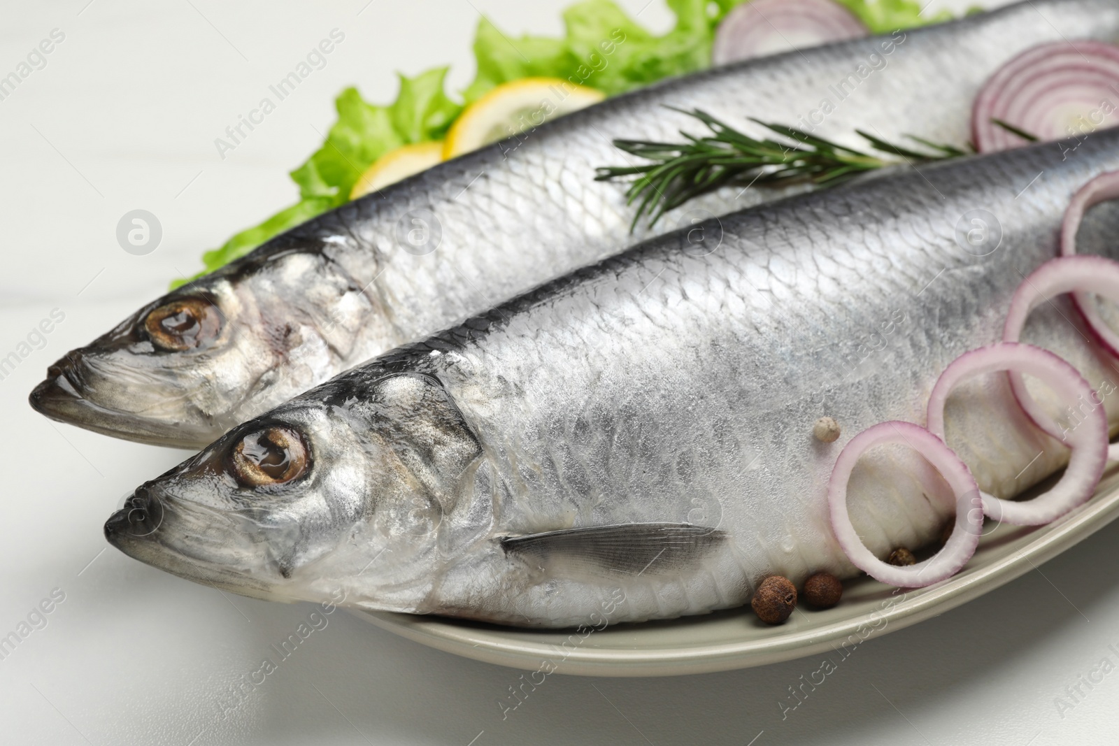 Photo of Plate with salted herrings, onion rings, slices of lemon, peppercorns and lettuce on white table, closeup