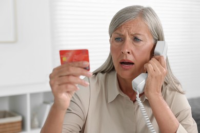 Photo of Worried woman with credit card talking on phone indoors. Be careful - fraud