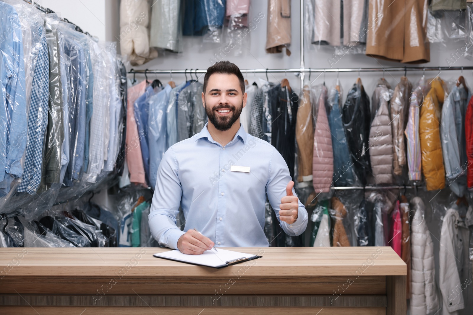 Photo of Dry-cleaning service. Happy worker showing thumb up at counter indoors