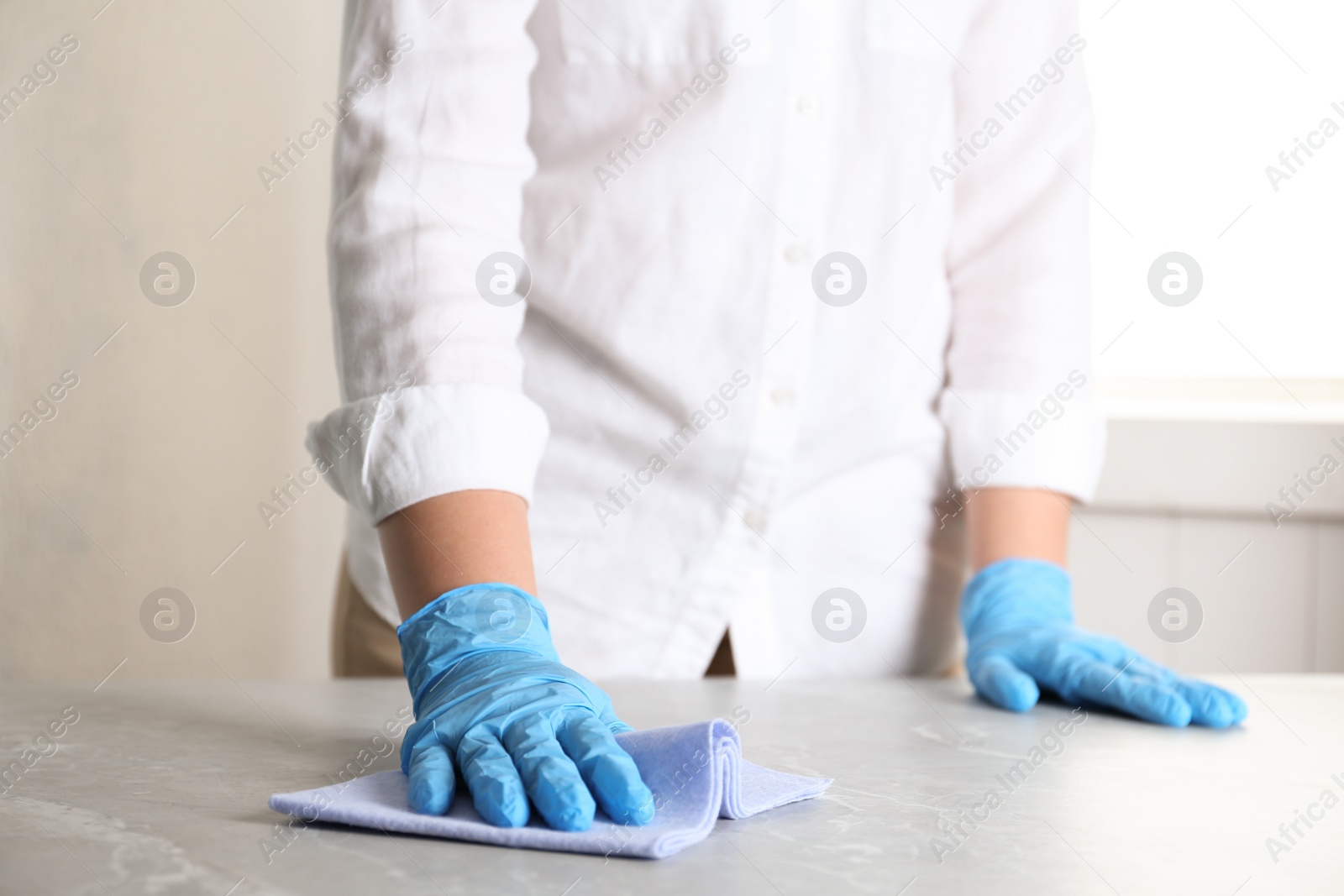 Photo of Woman in gloves wiping grey marble table with rag indoors, closeup