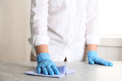 Photo of Woman in gloves wiping grey marble table with rag indoors, closeup