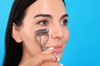 Beautiful young woman using eyelash curler on light blue background