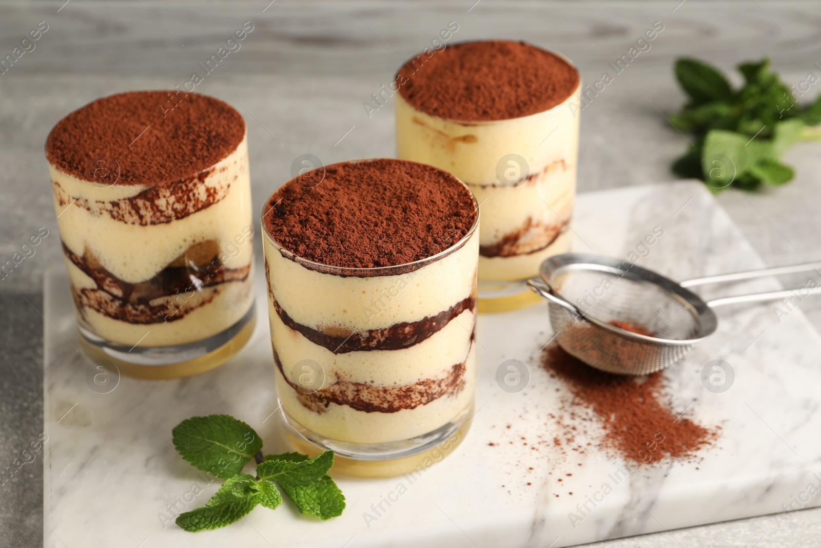 Photo of Delicious tiramisu in glasses, mint and sieve with cocoa powder on table, closeup