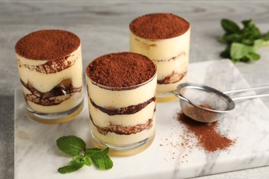 Photo of Delicious tiramisu in glasses, mint and sieve with cocoa powder on table, closeup