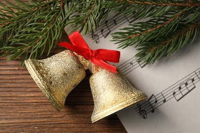 Photo of Golden shiny bells with red bow, music sheet and fir branches on wooden table, flat lay. Christmas decoration