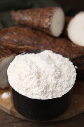 Photo of Scoop with cassava flour and roots on wooden table, closeup