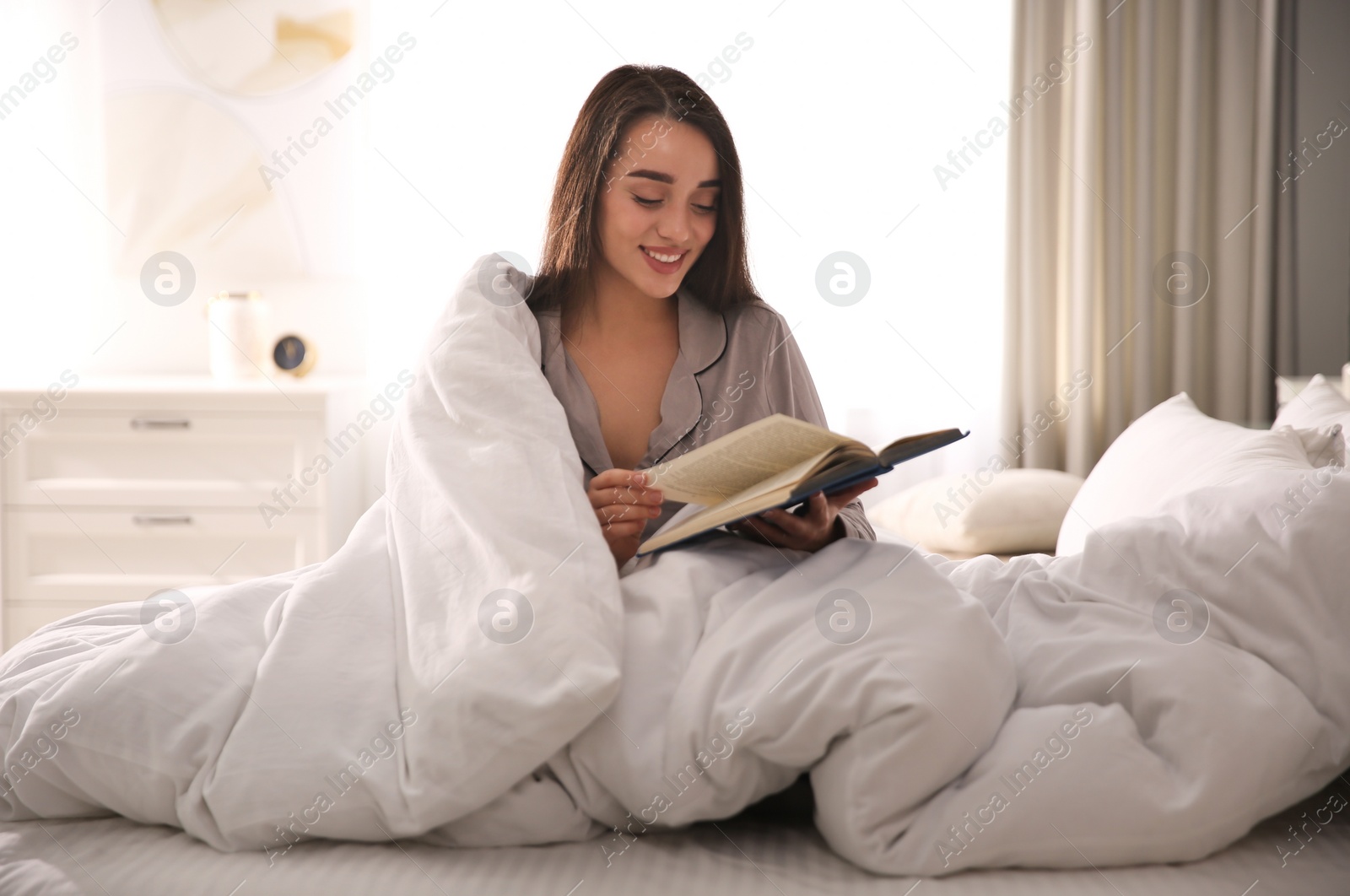 Photo of Beautiful young woman wrapped with soft blanket reading book on bed at home