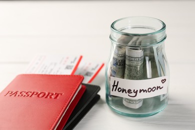 Glass jar with word Honeymoon, dollar banknotes, passports and plane tickets on white wooden table, closeup