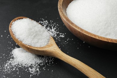 Photo of Granulated sugar in bowl and spoon on black table, closeup