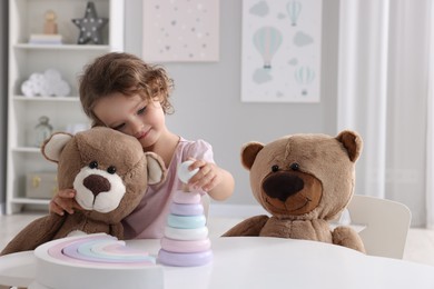 Photo of Cute little girl playing with toy and teddy bears at white table in room