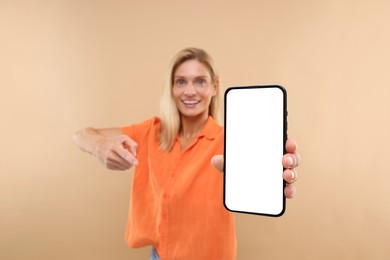 Happy woman holding smartphone and pointing at blank screen on beige background, selective focus