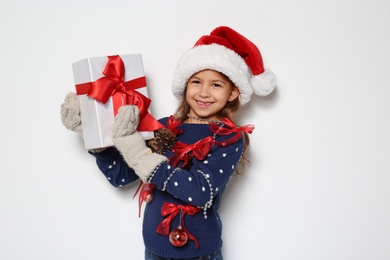 Cute little girl in handmade Christmas sweater and hat holding gift on white background