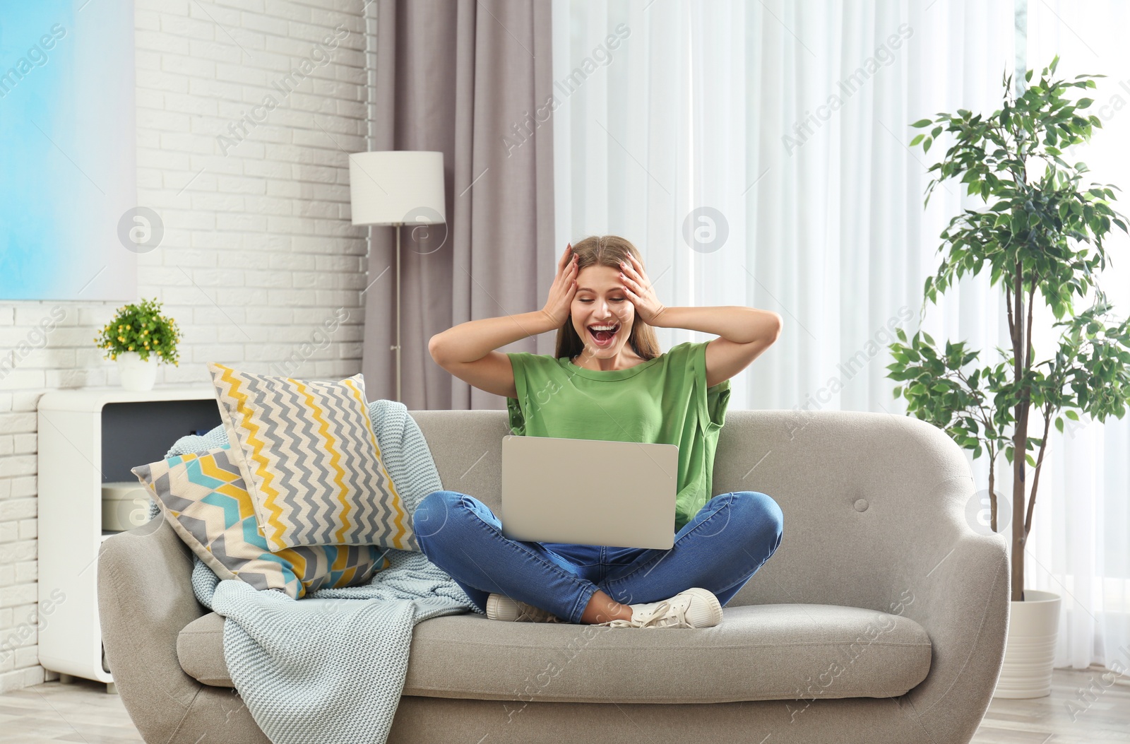 Photo of Emotional young woman with laptop celebrating victory on sofa at home