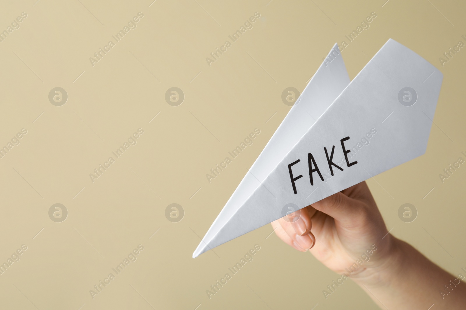 Photo of Woman holding paper plane with word FAKE near beige wall, closeup. Space for text