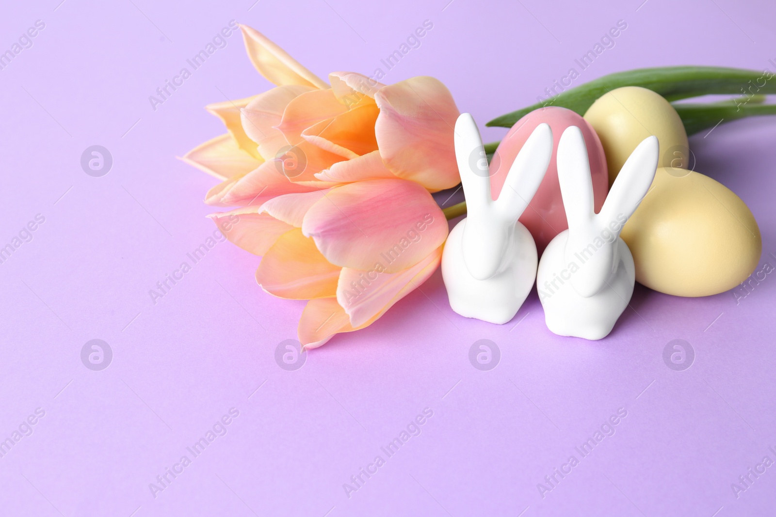 Photo of Cute ceramic Easter bunnies, dyed eggs and spring flowers on color background