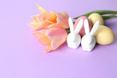 Cute ceramic Easter bunnies, dyed eggs and spring flowers on color background