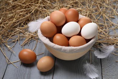 Fresh chicken eggs in bowl, feathers and dried straw on grey wooden table