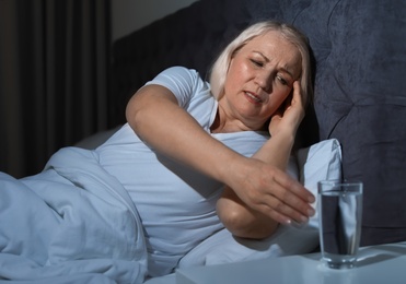 Photo of Mature woman with terrible headache at night taking glass of water from stand