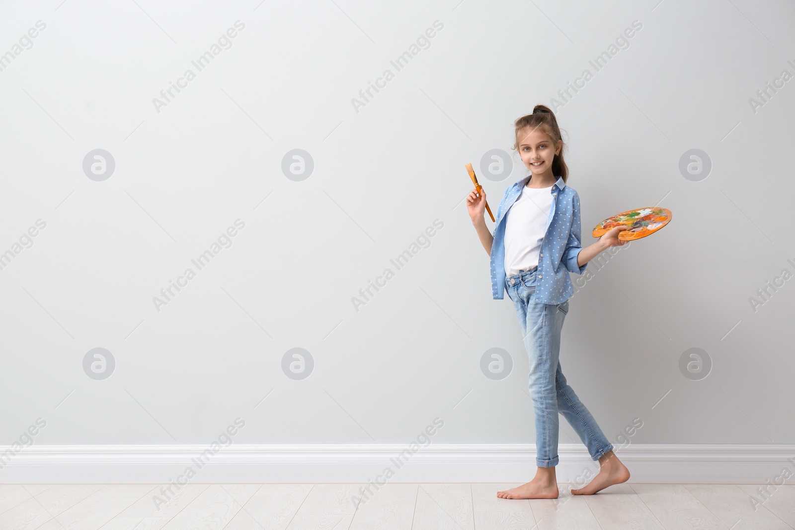 Photo of Little girl with brush and paint palette near light wall indoors. Space for text