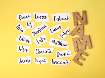 Photo of Paper sheets with written different baby names and wooden letters on yellow background, flat lay
