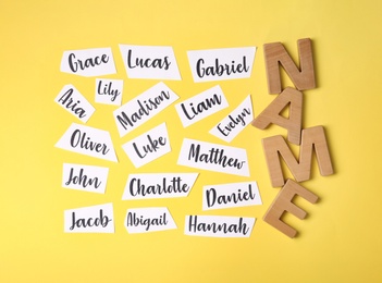 Paper sheets with written different baby names and wooden letters on yellow background, flat lay