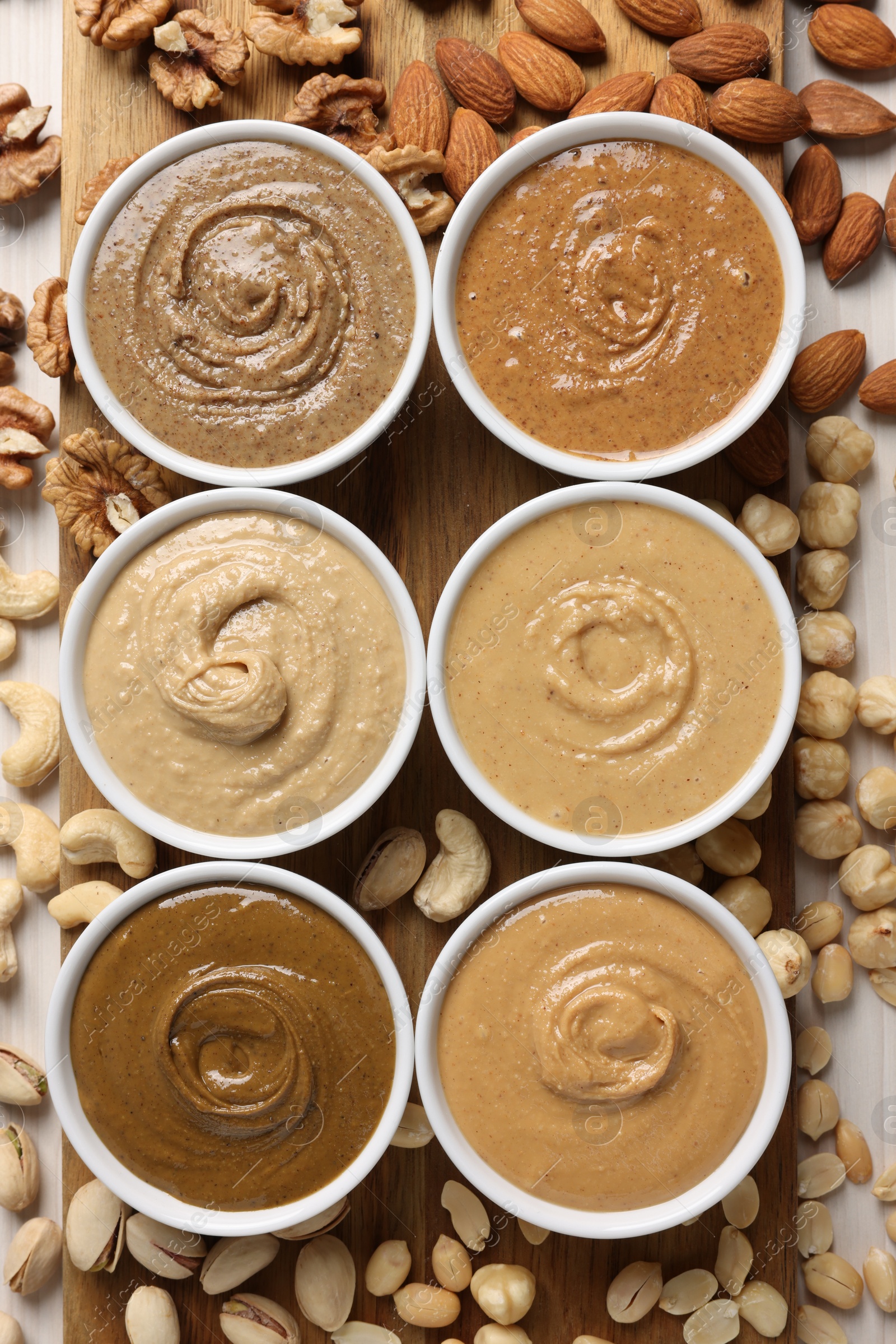 Photo of Many tasty nut butters in bowls and nuts on white table, flat lay