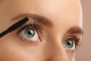 Woman applying mascara onto eyelashes against light brown background, closeup