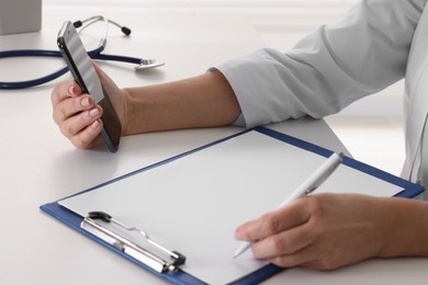 Photo of Doctor with smartphone and clipboard at desk indoors, closeup. Online medicine concept