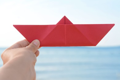 Child holding red paper boat near sea, closeup