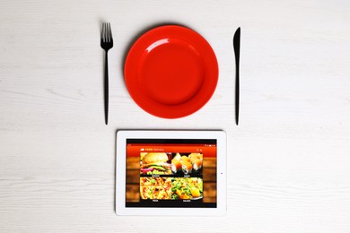 Photo of Modern tablet with open page for online food ordering, plate and cutlery on white wooden table, flat lay. Concept of delivery service