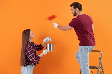 Photo of Man painting orange wall and happy woman holding can of paint. Interior design