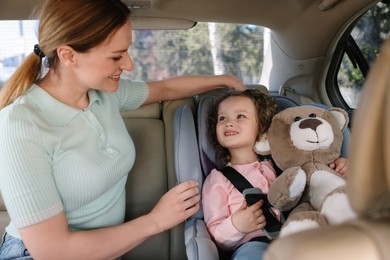 Photo of Cute little girl with teddy bear sitting in child safety seat near mother inside car
