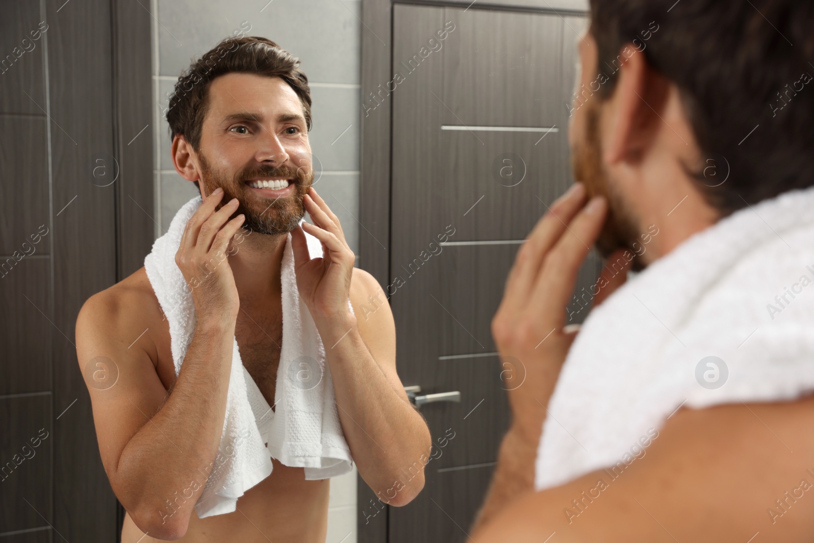 Photo of Handsome bearded man looking at mirror in bathroom near wooden doors