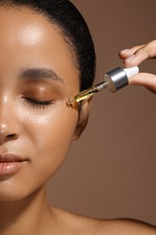 Woman applying serum onto her face on brown background, closeup