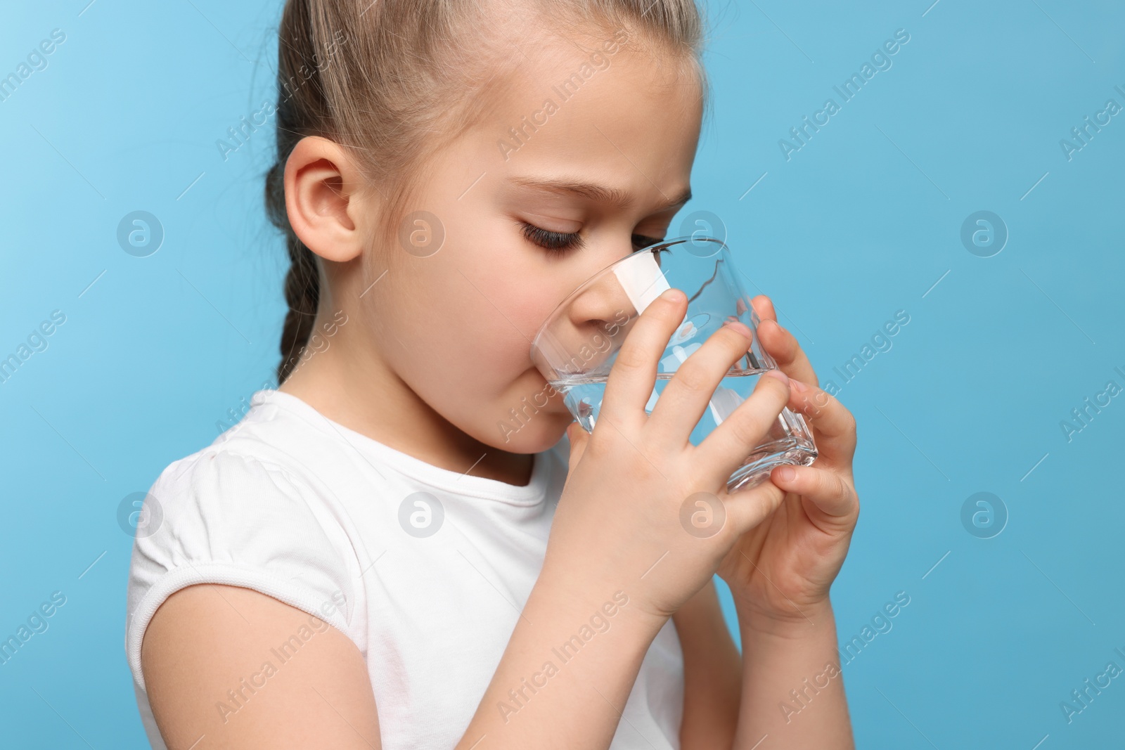 Photo of Cute little girl drinking fresh water from glass on light blue background