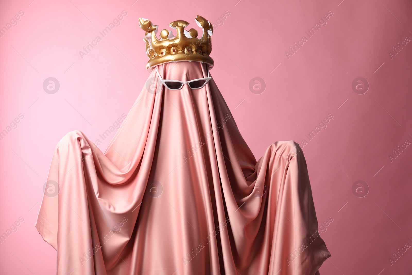 Photo of Glamorous ghost. Woman in sheet with sunglasses and crown on pink background