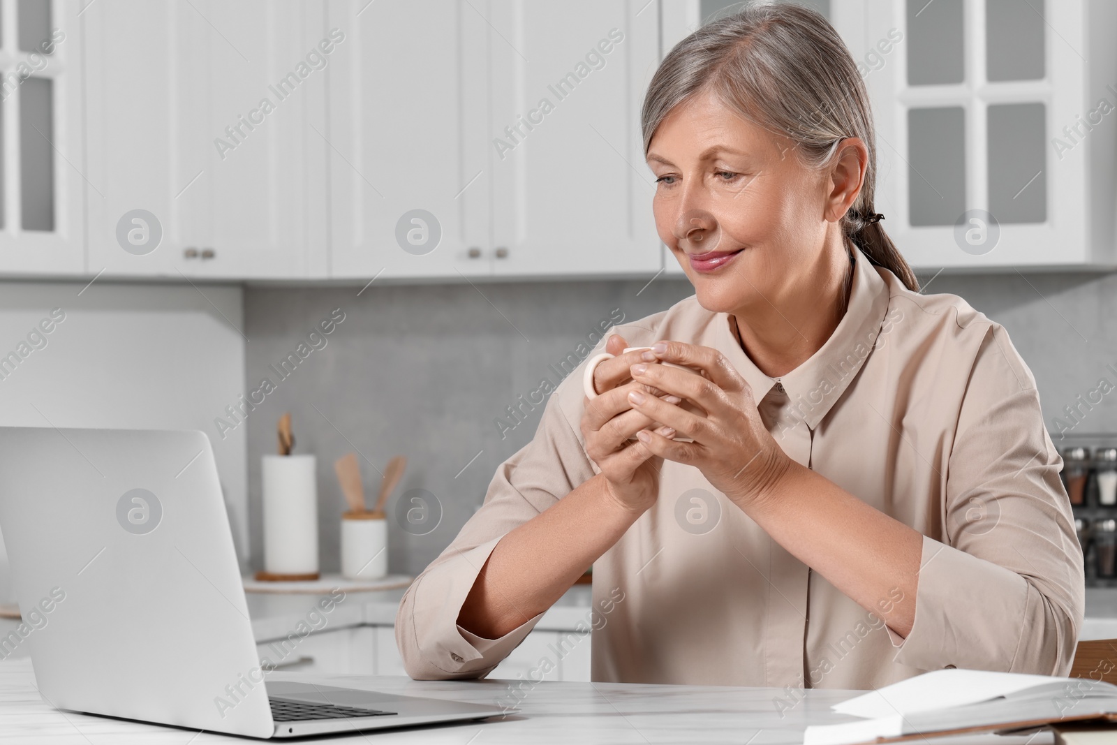 Photo of Beautiful senior woman with cup of drink near laptop at white marble table in kitchen