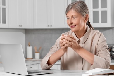 Photo of Beautiful senior woman with cup of drink near laptop at white marble table in kitchen