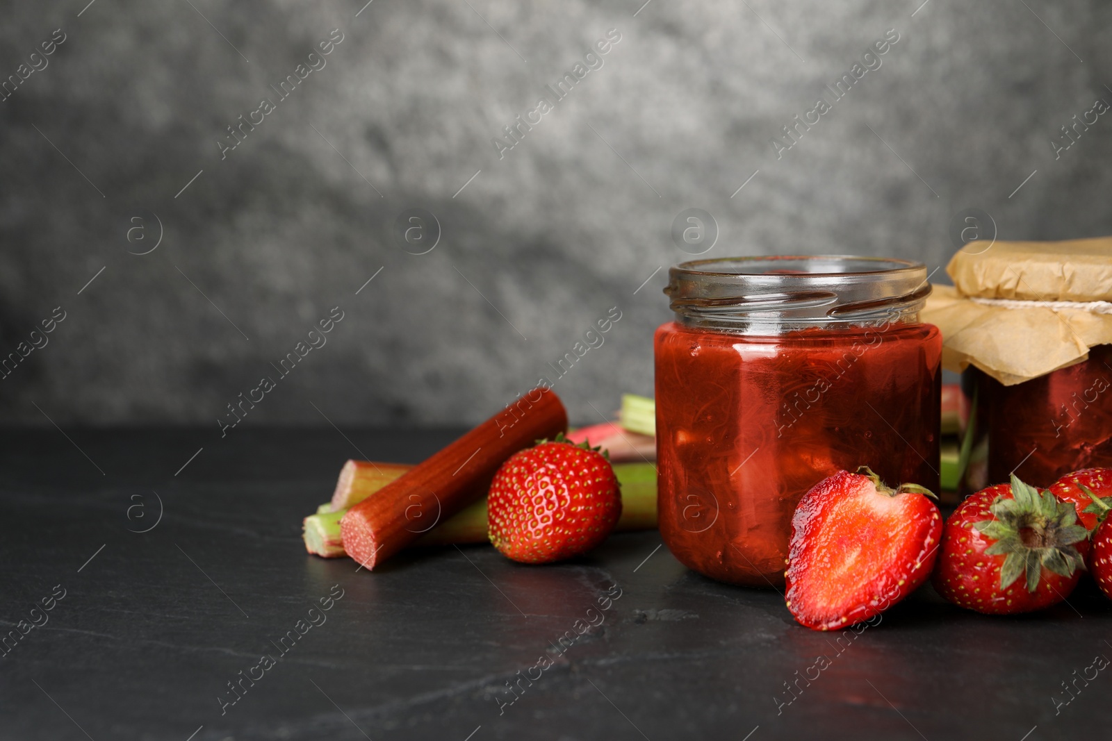 Photo of Jars of tasty rhubarb jam, fresh stems and strawberries on dark textured table. Space for text