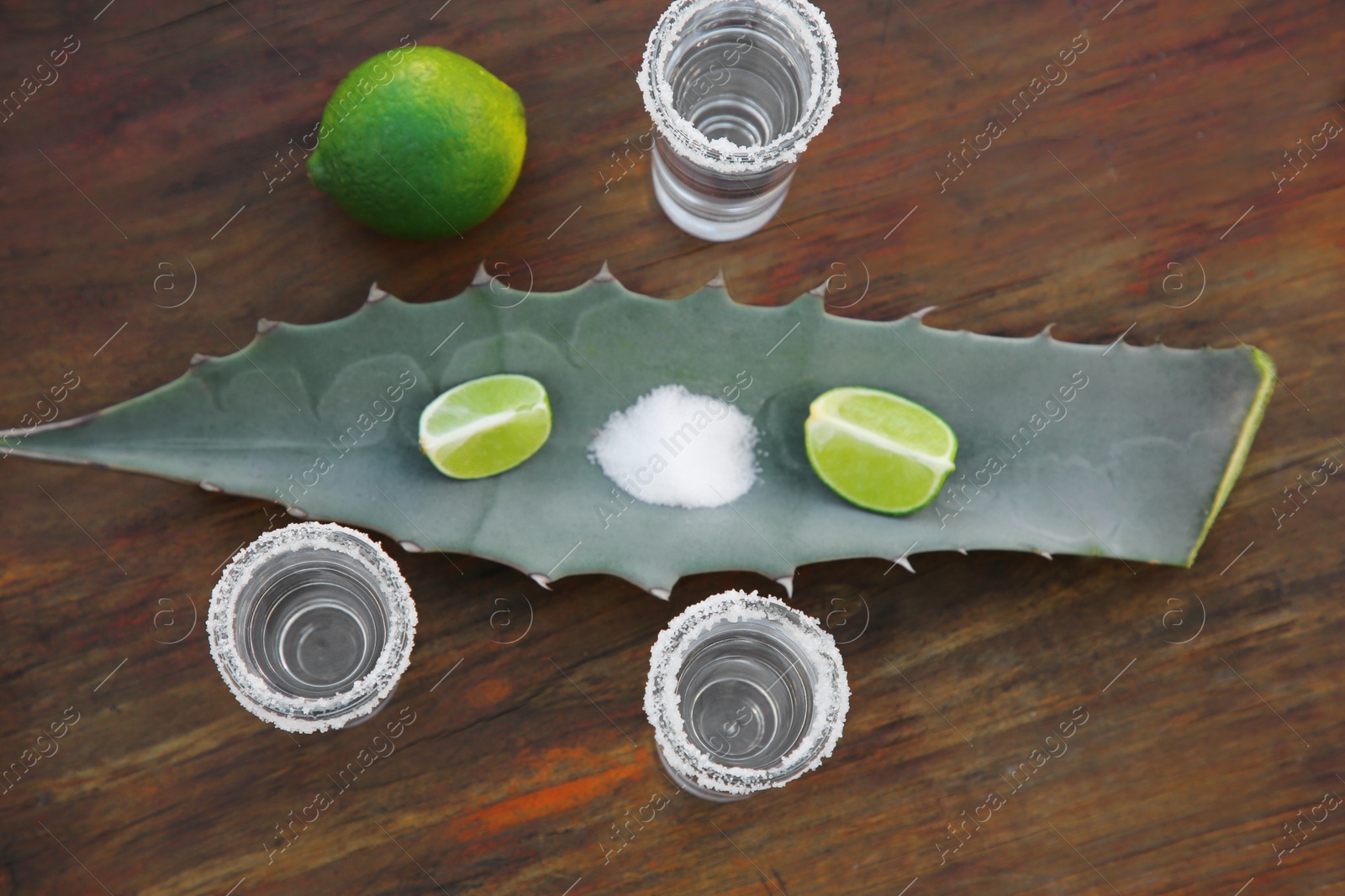 Photo of Mexican tequila shots, salt, lime and green leaf on wooden table, flat lay. Drink made of agava